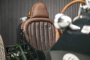 Close-up of a vintage car interior showing a brown leather seat with vertical stitching. The exterior of the car is dark green with a wooden steering wheel partially visible. The background features a dark corrugated surface.