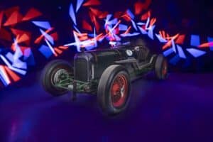 A vintage racing car with red and green colors is displayed against a dark background. Bright, abstract red and blue light trails create dynamic patterns surrounding the car, adding a sense of motion and energy to the scene.
