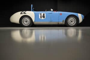 A classic blue and white vintage racing car with the number 14 on its side, parked in a sleek, modern setting. The car's design is simple and streamlined, with reflective flooring highlighting its silhouette.