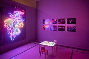 A room lit with purple neon light features a large glowing neon phoenix on the wall, a small table with two chairs, and colorful photos displayed on the adjacent wall.