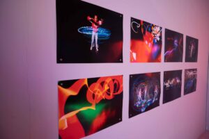 Eight colorful, abstract light painting photographs are displayed on a pale purple wall. The images feature swirling, vibrant light trails and some include people interacting with the lights.