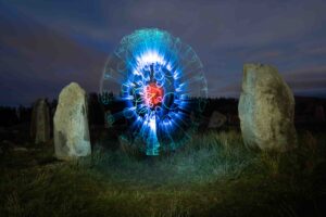 A glowing blue and red circular light pattern appears in the center of a grassy field surrounded by standing stones at night, creating a surreal and mystical effect.