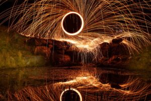 Long-exposure photo of swirling orange sparks creating circular patterns in the night sky, with rocks and water below reflecting the bright light trails.