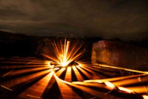 Long-exposure photo of spinning sparks creating bright, orange light trails in a circular pattern at night, with dark clouds and large rocks in the background.