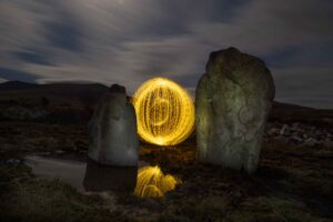 A glowing yellow light orb stands between two large rocks at night, reflected in a small puddle below. Dark hills and a cloudy sky are visible in the background.