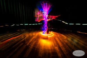 A long-exposure photo captures swirling, colorful lights forming a vibrant spiral vortex above a wooden floor in a dark room. The effect creates dynamic movement and bright, neon-like trails.