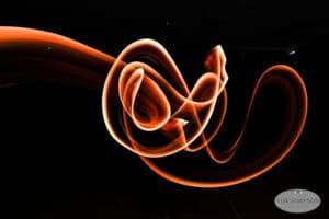 Swirling streaks of orange and white light form abstract shapes against a black background. The light trails curve and twist dynamically, creating a sense of motion. The photographer’s watermark is visible in the bottom right corner.