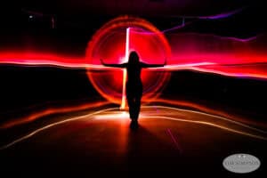 A person stands with arms outstretched in front of swirling red and orange light trails, creating a dramatic glowing halo effect in a dark room. The scene is artistic and striking.