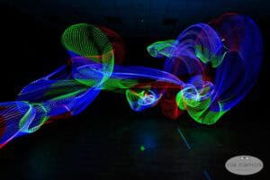 A dark room is illuminated by swirling trails of blue, green, and red lights, creating abstract patterns in the air. The floor is dimly visible, and the image has a watermark that reads "Tim Simpson Photography.