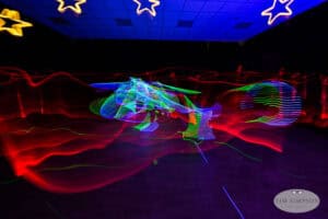 A dark room with glowing neon light trails in red, green, and blue, creating abstract patterns. Yellow star decorations hang from the ceiling, and the photo has a Tim Simpson Photography watermark.