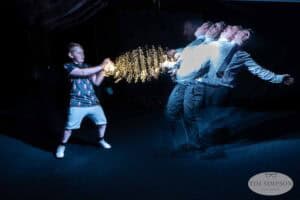 A boy in shorts and a printed shirt pretends to shoot sparking lights from his hands at three men in suits, who stagger backward in a dramatic, motion-blur effect. The scene is dark with a playful, staged atmosphere.
