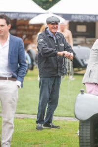 An older man in a flat cap and jacket smiles while pouring water from a bottle onto the grass at an outdoor event, with other people and classic cars in the background.