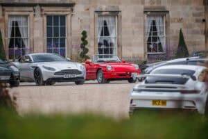 Several classic and modern sports cars are parked on a gravel driveway in front of a historic stone building with large windows and topiary trees. A bright red convertible stands out among the vehicles.