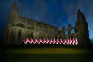 Ruins of a large stone building at night, illuminated by a dark blue sky, with a wavy pattern of red and white light trailing across the grass in front of the structure.
