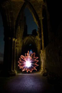 A long-exposure photo shows a glowing, spinning light pattern resembling a flower inside a dark, ancient stone ruin with tall arches.