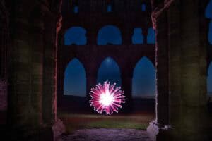 A bright, glowing orb with pink and white light trails stands in the center of ancient stone ruins at night, framed by large arches and columns against a dark blue sky.