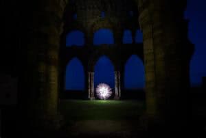 A circular light pattern glows in the center of ancient stone arches at night, illuminating the ruins against a deep blue sky.