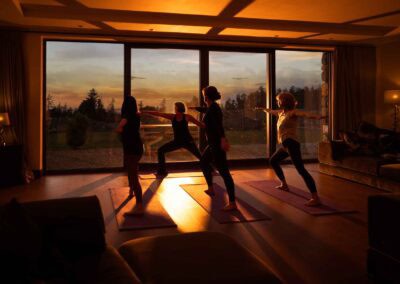 Four people practice yoga indoors at sunset, silhouetted against large windows with an outdoor view. Warm light fills the room as they stand on yoga mats in a warrior pose, creating a serene scene perfect for photographing a writing retreat.
