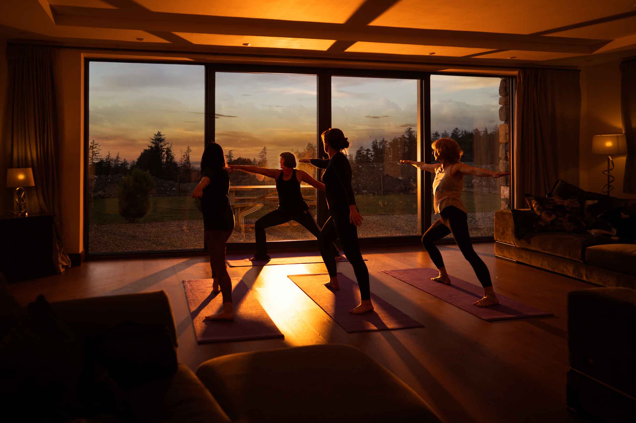 yoga photography Four people practice yoga indoors at sunset, silhouetted against large windows with an outdoor view. Warm light fills the room as they stand on yoga mats in a warrior pose, creating a serene scene perfect for photographing a writing retreat.