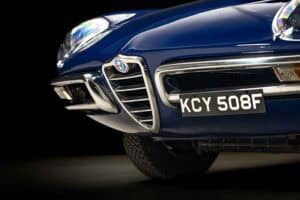 Close-up of the front grille and bumper of a classic blue Alfa Romeo car, showing the Alfa Romeo badge and a license plate reading "KCY 508F" against a dark background.