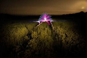 A field of tall grass at night with colorful light trails forming a fan-like pattern in the distance under a cloudy, orange-tinted sky.