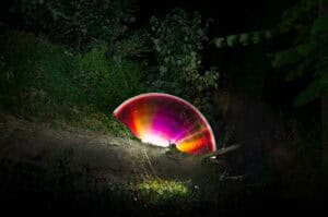 A vibrant, multicolored semicircle of light glows behind a fallen tree in a dark, wooded area, illuminating nearby grass and foliage.
