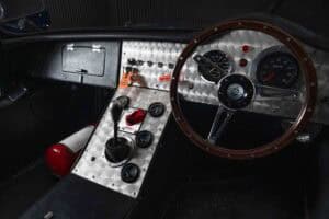 Close-up of a classic car interior showing a wooden steering wheel, gauges, metal dashboard with toggle switches, gear shift, and a red fire extinguisher on the floor.