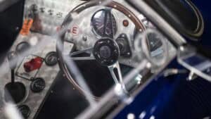 Close-up view of a vintage race car’s cockpit showing a wooden steering wheel, dashboard with gauges and switches, and a manual gear shift lever. The image captures metallic details and reflective surfaces.