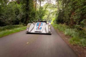 A silver and blue race car with a driver speeds down a narrow, tree-lined road, surrounded by lush green foliage on both sides.
