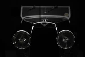Close-up of the front of a vintage car with two round headlights, a chrome hood ornament, and a windshield in the background, set against a dark, shadowy background.