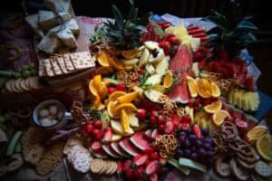 A vibrant charcuterie board displays assorted fruits like watermelon, strawberries, oranges, pineapple, grapes, cheeses, crackers, pretzels, and two decorative pineapples on a colorful tablecloth.