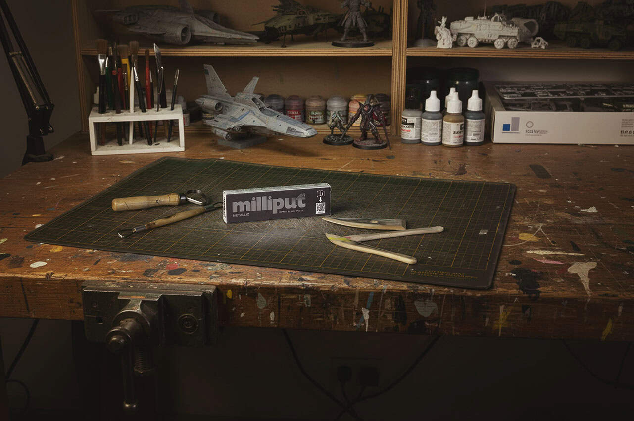 A workbench with modeling tools, a cutting mat, sculpting tools, and a box of Milliput. Paintbrushes, paints, and assembled model vehicles and figures are on shelves in the background.