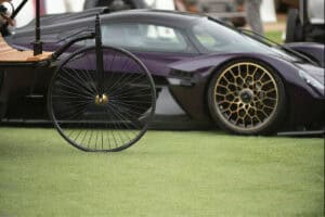 A close-up of a vintage bicycle wheel in the foreground, with a modern black supercar featuring gold rims parked on grass in the background.