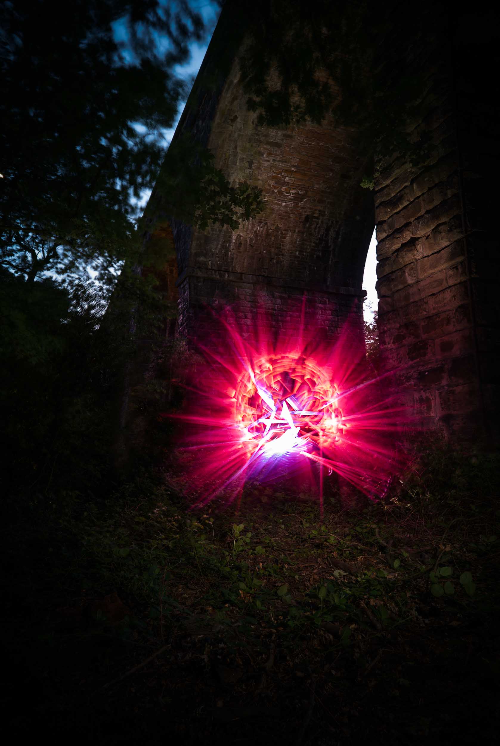 A stone bridge stands in the dark as a glowing, bright pink and purple circular light effect radiates beneath its arch, illuminating the surrounding foliage and ground.