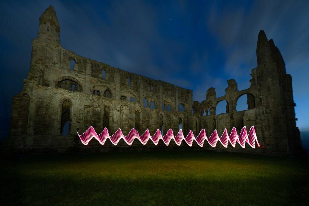 Ruins Whitby Abbey at night, with swirling red and white light trails in front of it, illuminating the grass and contrasting against the dark blue sky.