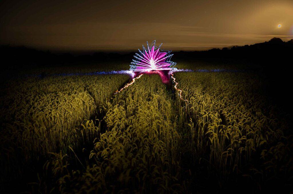 A nighttime photo of a field with tall grass and tractor tracks, featuring vibrant, fan-shaped light trails in pink, purple, and blue, illuminating the center under a cloudy sky with a glowing moon.
