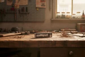 A box of Milliput epoxy putty sits on a wooden workbench surrounded by metal shavings and tools, with jars and equipment visible in the background of a sunlit workshop.