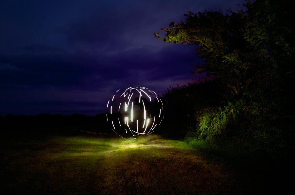 A glowing orb created with white light trails floats above grass at night, surrounded by dark trees and bushes under a deep blue and purple sky.