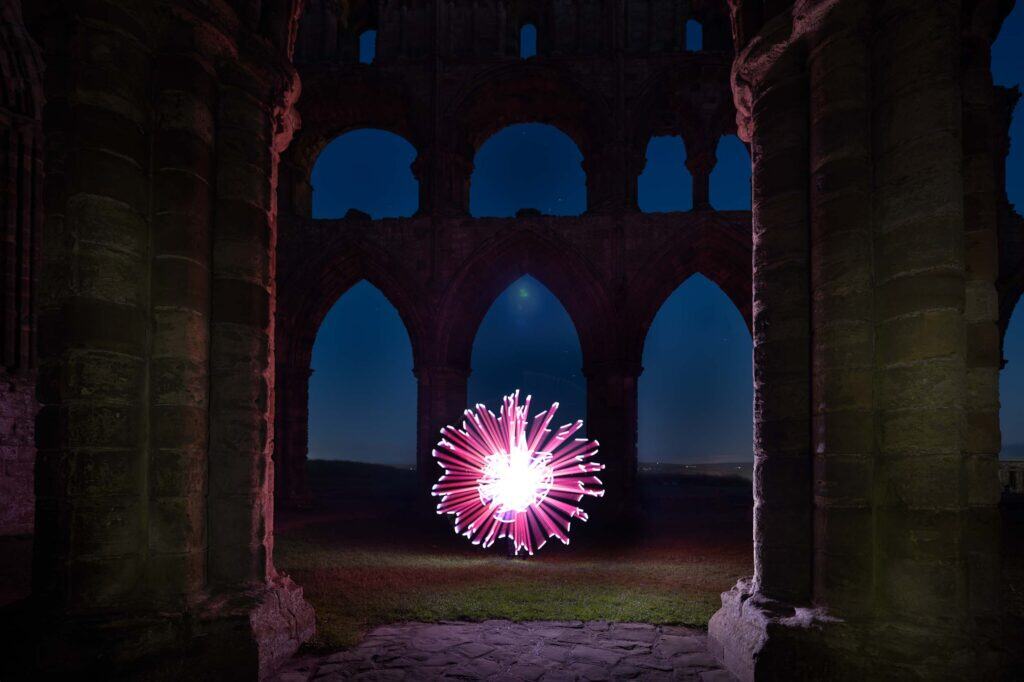 A glowing pink and white light orb is centered between ancient stone arches of Whitby Abbey at night, with dark sky and ruins silhouetted in the background. The scene is framed by two large stone columns in the foreground.
