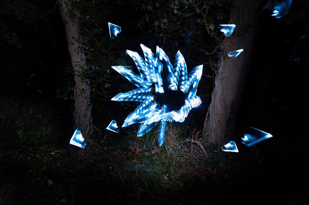 Bright blue and white light trails form a spiky, abstract shape between two tree trunks in a dark, wooded area, with smaller fragments floating nearby.