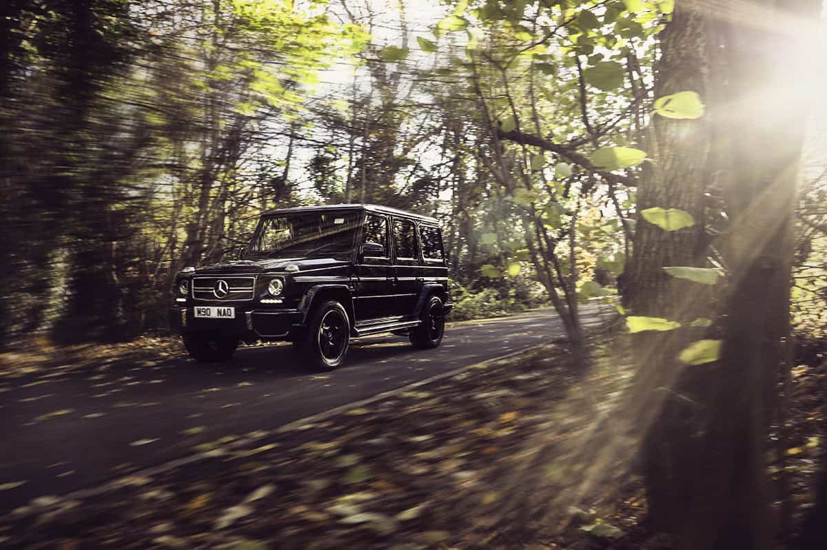 A black Mercedes-Benz G-Class SUV drives down a sunlit, tree-lined road in a forest. Sunbeams filter through the autumn leaves, creating a warm, golden atmosphere.