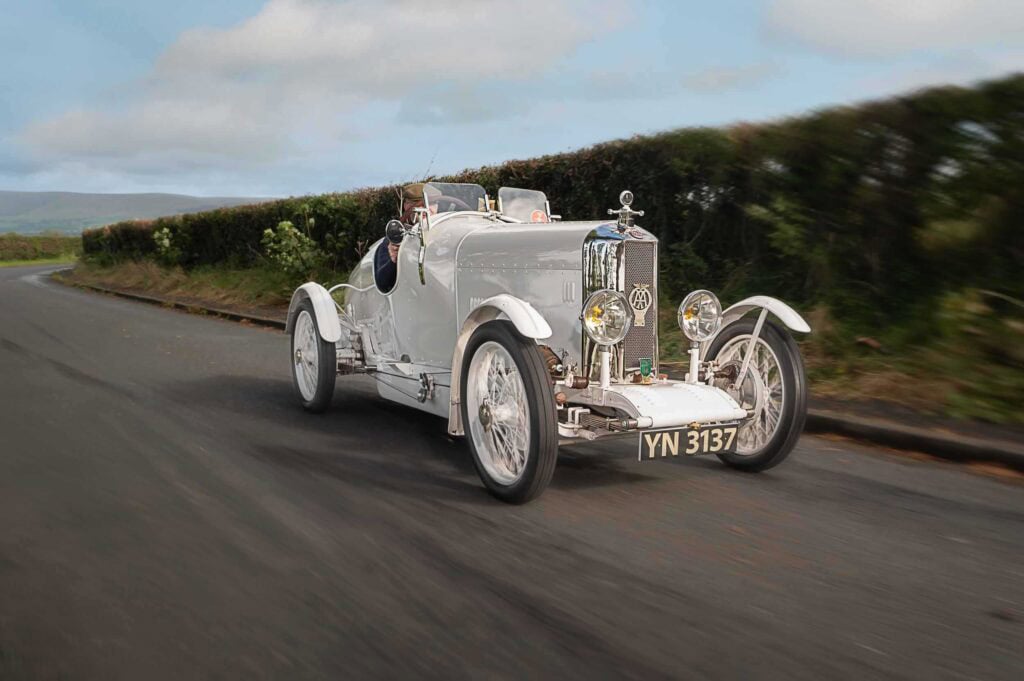 A classic Rally grans sport S vintage car with the license plate YN 3137 drives on a country road, bordered by green hedges under a partly cloudy sky.