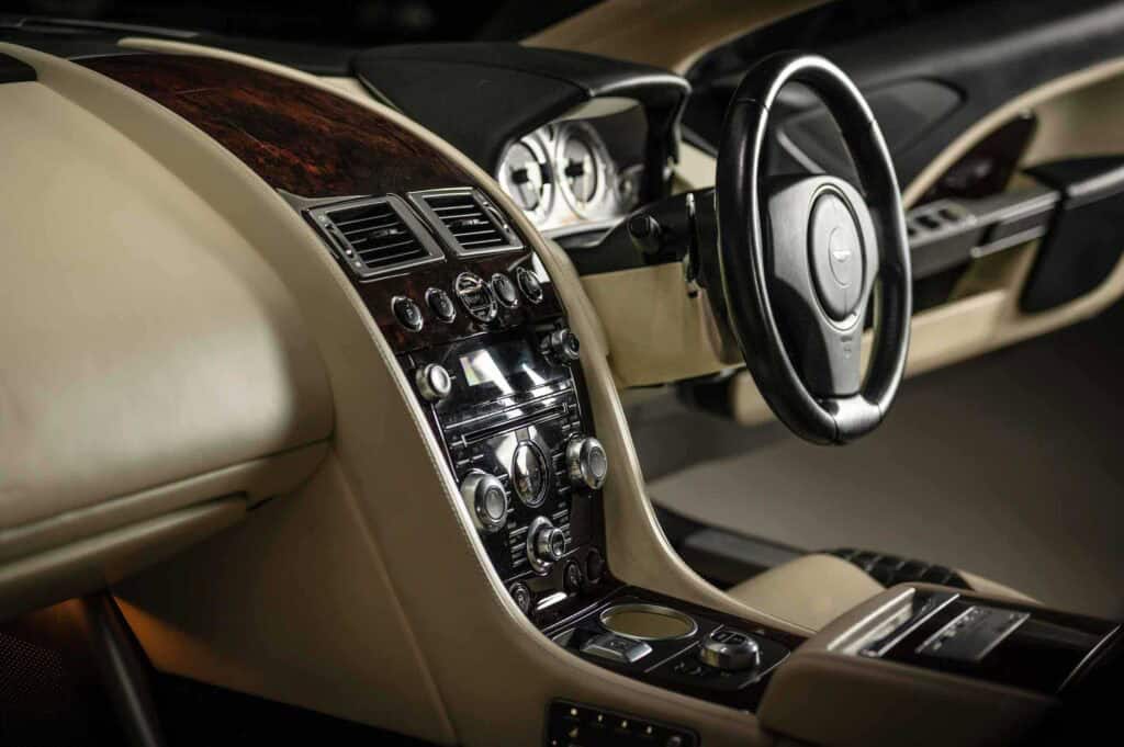 Aston Martin car interior featuring a right-hand drive steering wheel, beige leather seats, polished wood trim, and a sleek central control panel with multiple dials and buttons.