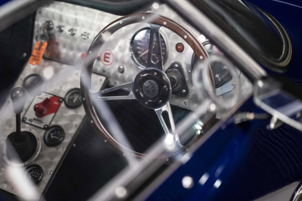 Close-up view of a marcos martin's interior, showing a metal steering wheel with wooden trim, dashboard gauges, switches, and gear stick through the window. The surfaces are metallic and the car’s exterior is blue.