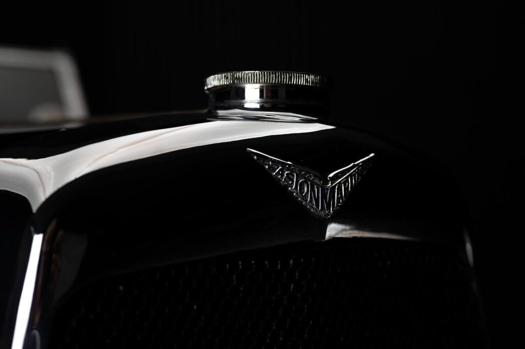 A close-up of a shiny vintage car hood with an Aston Martin emblem and a metal radiator cap, set against a dark background.
