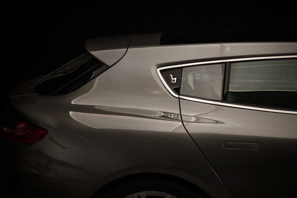 A close-up side view of the rear section of an astir martin shooting brake designed by Bertone highlighting the smooth metallic surface, sharp lines, rear window, and a small badge. The background is dark, making the car stand out. it shows the Bertone logo.