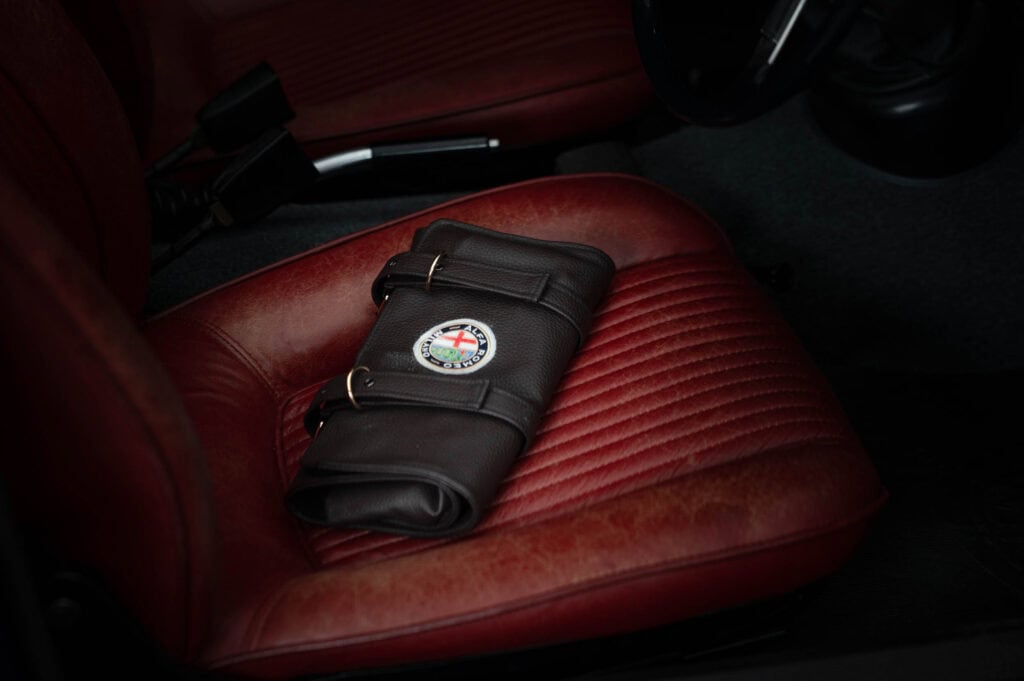 A leather alfa romeo leather owners manual case with a colourful circular badge rests on the driver’s seat of a car with red leather upholstery. The car’s gear stick and steering wheel are partially visible.