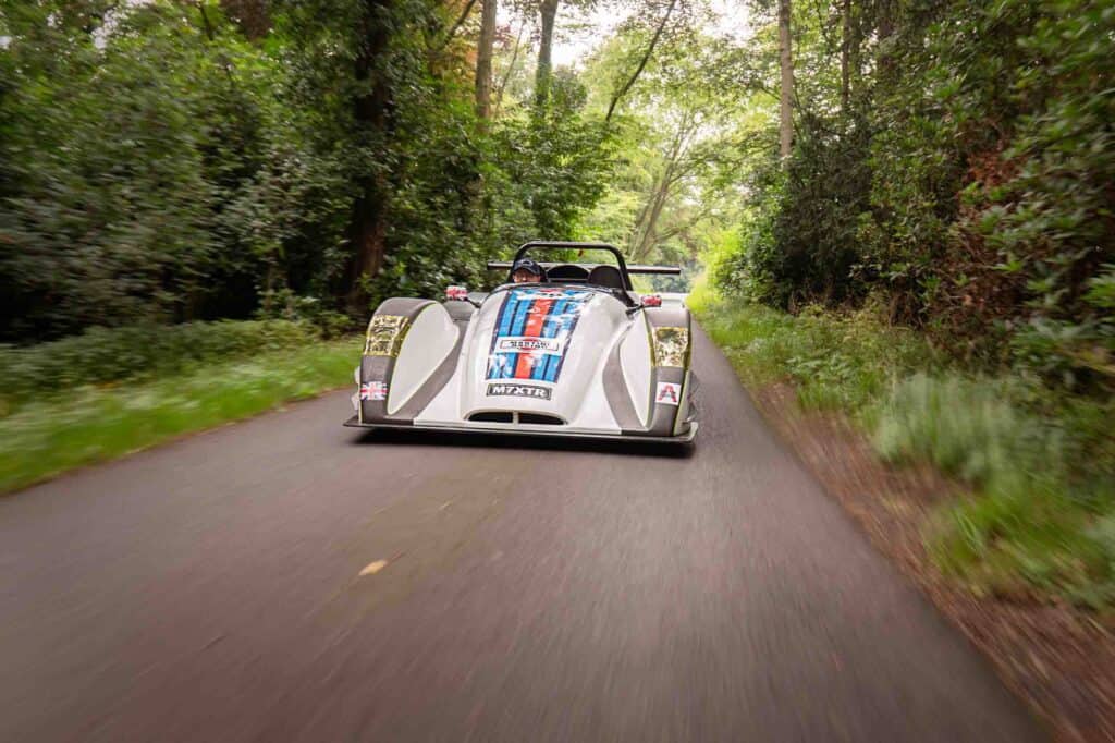 A westfield blue and red martini stripes drives quickly down a narrow, tree-lined road, surrounded by lush green foliage. The image captures a sense of speed and motion.