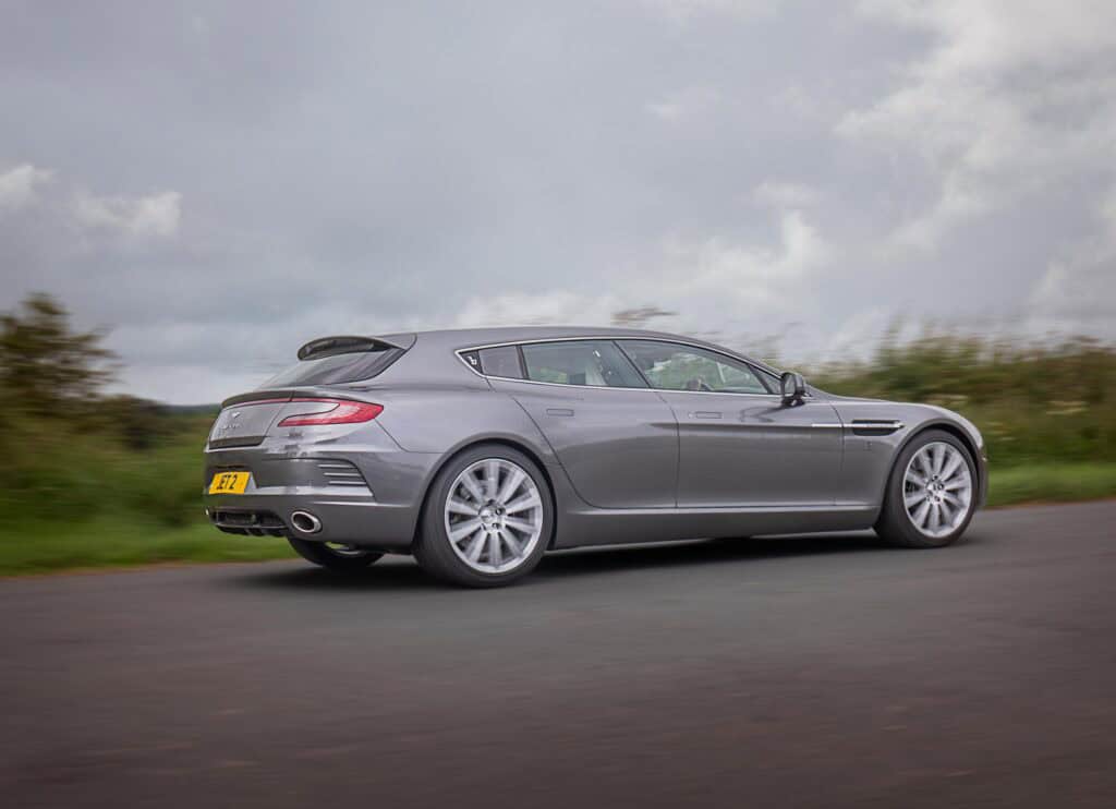 Aston Martin Jet 2 shooting brake with four doors is driving on a country road, surrounded by greenery and an overcast sky. The car is viewed from the rear side, emphasizing its sporty, elegant design.