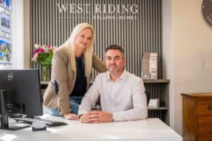 A woman stands beside a seated man at a desk in a modern office. Both are smiling. The sign behind them reads “West Riding Selling Homes.” There’s a computer, flowers, and property brochures visible on the desk.